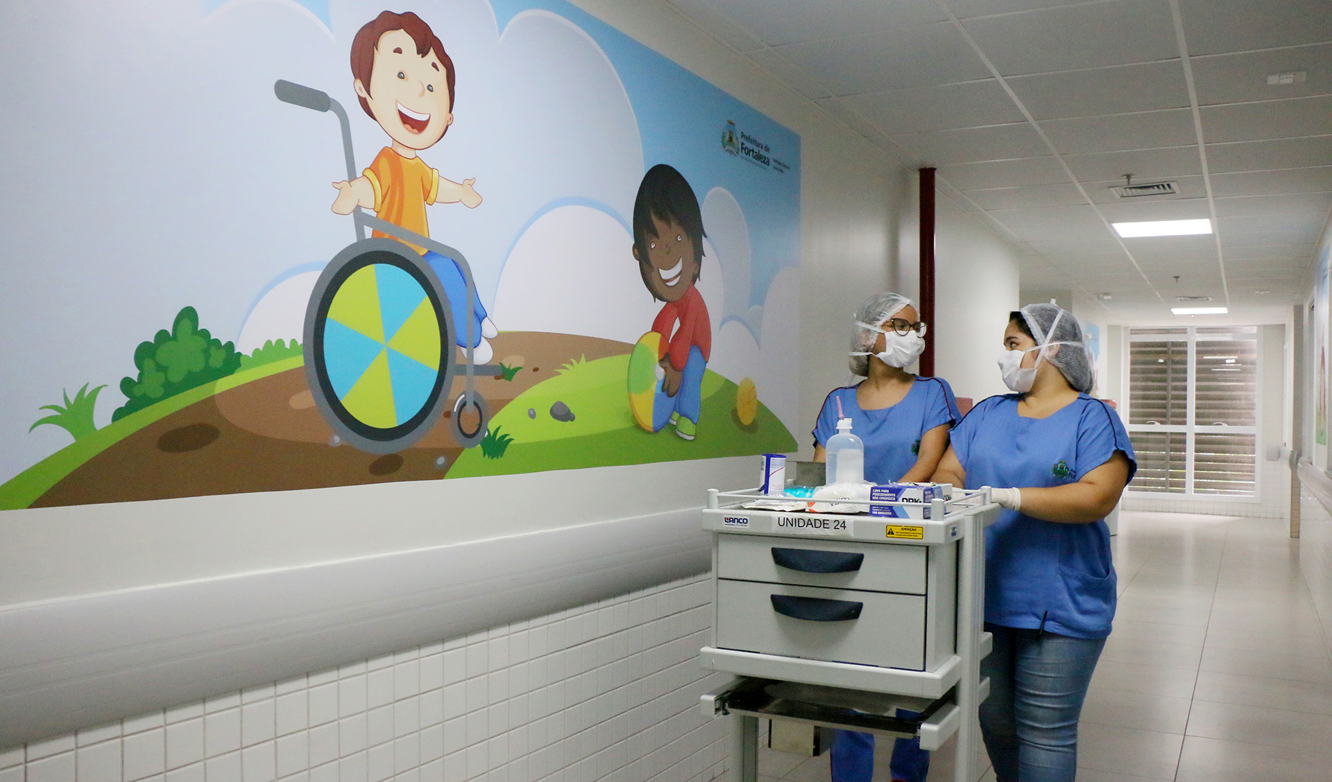 duas moças com roupa, touca e máscara de enfermeira andando em corredor empurrando carrinho com materiais